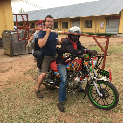 Kyle Reeser transporting a metal rack on a motorcycle at LVDC.  The Lake Victoria Disability Centre Campus consisted of many yellow buildings with corrugated metal roofs, all built and maintained by LVDC staff.  Lake Victoria Disability Centre | Musoma, Tanzania.